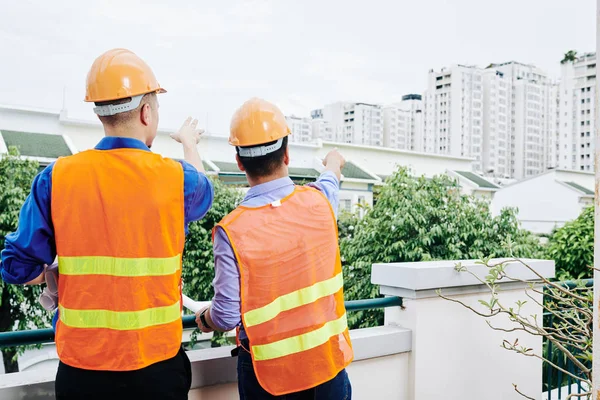 Widok Tyły Inżynierów Pomarańczowych Kamizelkach Wskazujących Budynek Nowej Dzielnicy Którą — Zdjęcie stockowe