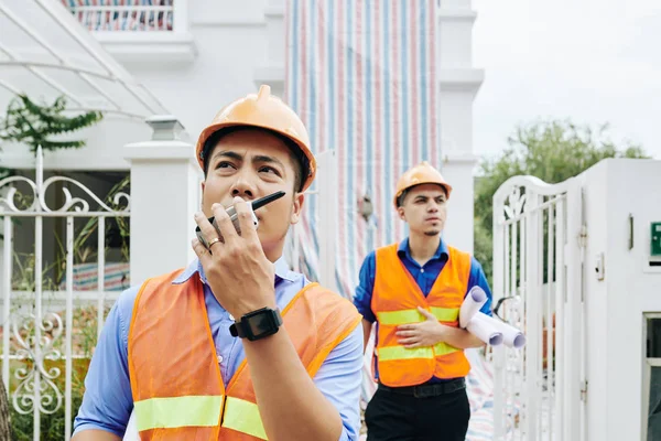 Serious frowning Vietnamese construction engineer talking on walkie-talkie when he and his coworker leaving building they were working in