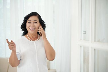Asyalı mutlu, olgun bir kadın cep telefonuyla konuşurken gülümsüyor ve evde dikilip işaret ediyor.
