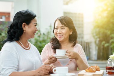 İki Asyalı gülümseyen kadın kahvelerini yudumlayıp açık kafede birbirleriyle konuşuyorlar.