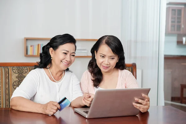 两个亚洲成熟的女人一起坐在桌旁 看着她们用信用卡在网上购物的笔记本电脑 — 图库照片