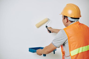 Duvarı boyarken boyayı yakalamak için tepsi kullanan turuncu yelek ve kasklı bir ressam.