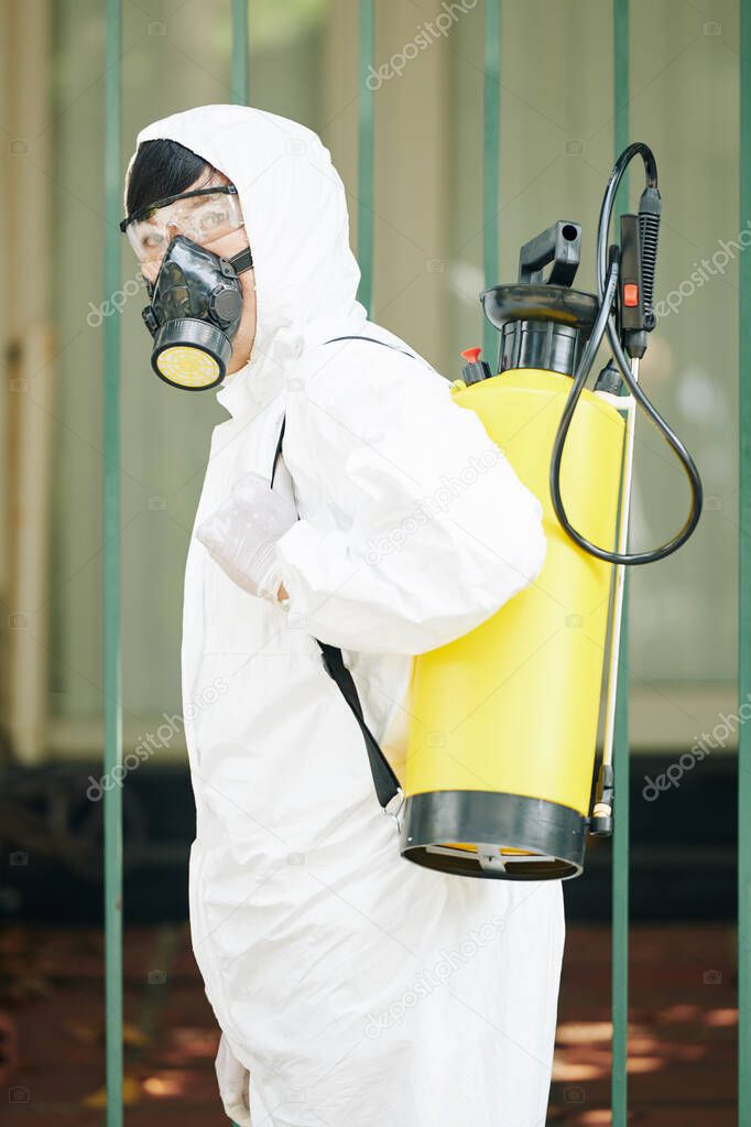 Portrait of Asian technician in a protective suit and mask spraying chemicals and turning back to look at camera