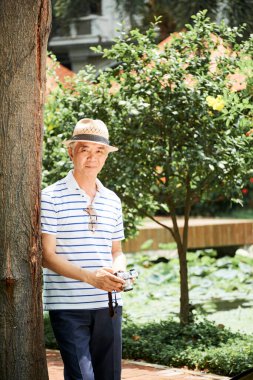 Gülümseyen yaşlı Çinli adam, şehir parkında ya da bahçede çiçek, ağaç ve doğa fotoğrafları çekmekten zevk alıyor.