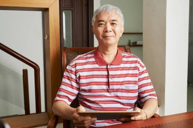 Çizgili tişörtlü mutlu Çinli adamın portresi. Evdeki masasında tablet bilgisayarda çalışıyor.