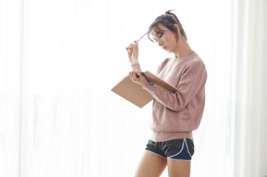 Düşünceli Vietnamlı bayan öğrenci kitabı okuyor ve kalemle alnını kaşıyor.