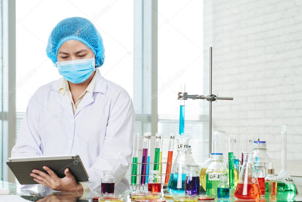 Young serious Asian female laboratory worker filling form on tablet computer after doing research in clinic