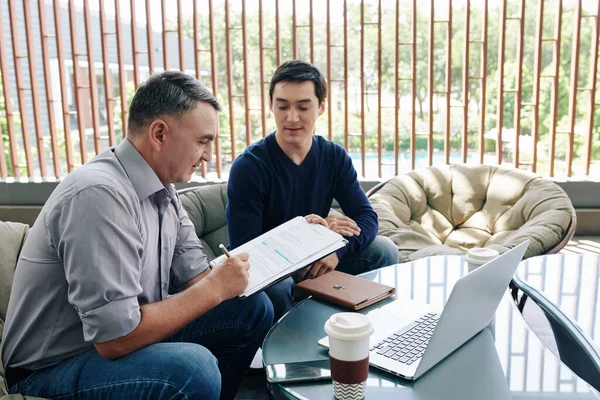 Mature entrepreneur signing contract with young software developer after having meeting in office lounge area