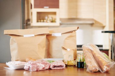 Mutfak masasında içinde yiyecek, et ve taze ekmek olan kağıt paketler.