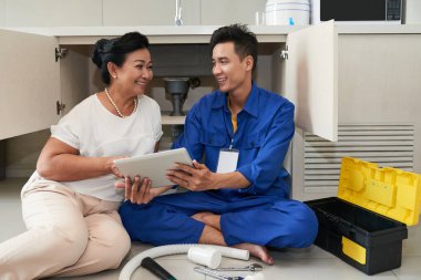 Gülümseyen tesisatçı yaşlı kadın tablet bilgisayarını gösteriyor ve mutfak giderlerini nasıl tamir edeceğini anlatıyor.
