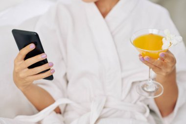 Kaplıca merkezinde akıllı telefon ve meyve kokteyliyle rahatlayan genç bir kadının kırpılmış görüntüsü.