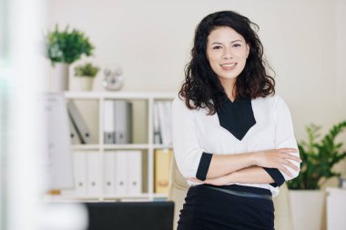 Ofis masasında kollarını kavuşturup kameraya bakan neşeli genç iş kadını portresi.