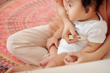 Anne hasta çocuğunun sıcaklığını elektrikli termometreyle kontrol ediyor.