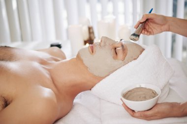 Güzellik salonunda dinlenen genç adamın yüzüne çamur maskesi takan güzellik uzmanının elleri.