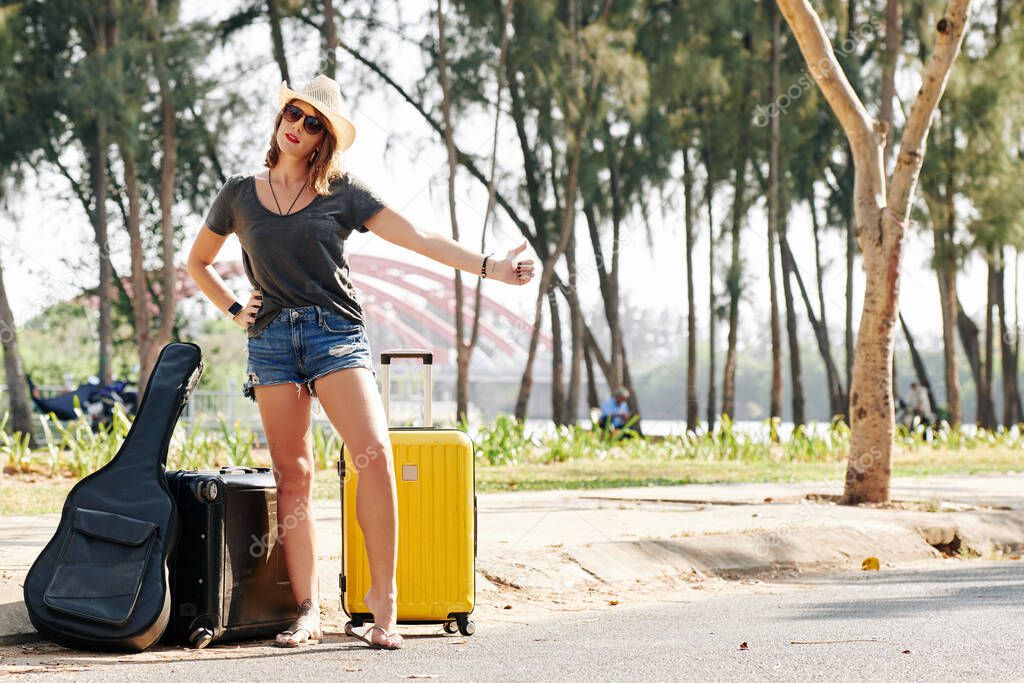 Slim pretty young woman in denim shorts standing on roadside with suitcases and guitar and catching car