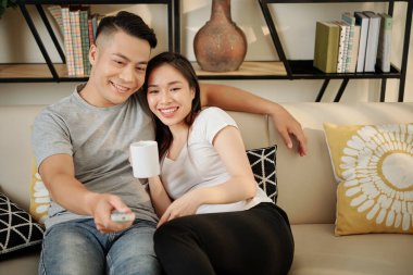 Mutlu, güzel, genç Vietnamlı kadın evde erkek arkadaşıyla televizyon izlerken bir fincan kahve içiyor.