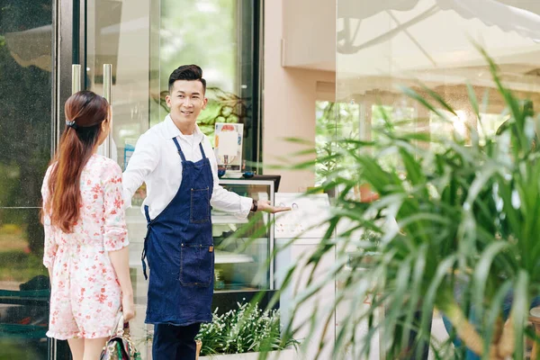 兴高采烈的年轻的越南咖啡馆店主 穿着牛仔围裙 欢迎女顾客来到他的咖啡馆 — 图库照片
