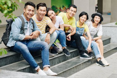 Üniversitenin ya da üniversite binasının merdivenlerinde oturmuş kameralara bakan neşeli Asyalı genç insanlar.