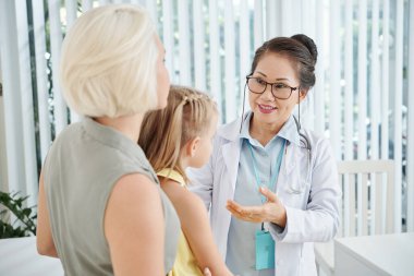 Gülümseyen deneyimli bir çocuk doktoru küçük bir kızın annesiyle konuşuyor ve reçeteyi açıklıyor.