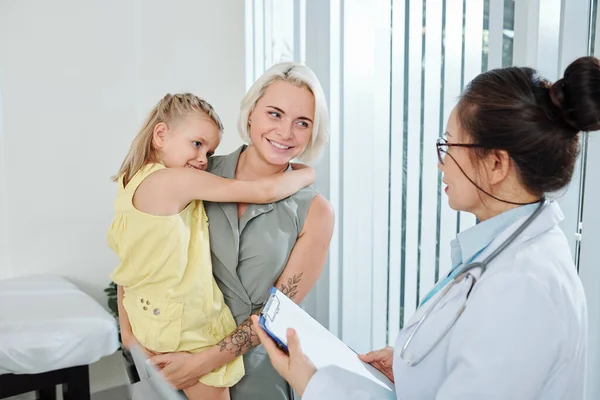 診断や処方について小児科医と話すとき 彼女の小さな娘を運ぶかなり笑顔若い女性 — ストック写真