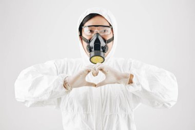 Koruyucu takım elbiseli, maskeli ve gözlüklü bir sağlık görevlisi ellerini kalp şeklinde tutuyor.