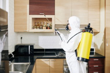 Anti-koronavirüs dezenfeksiyon servisi görevlisi. Tehlikeli madde giysisi içinde. Müşterinin mutfağındaki tüm yüzeylere sprey sıkıyor.