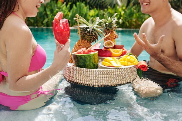 Cropped image of young couple sitting in swimming pool and eating tasty fresh fruits for breakfast