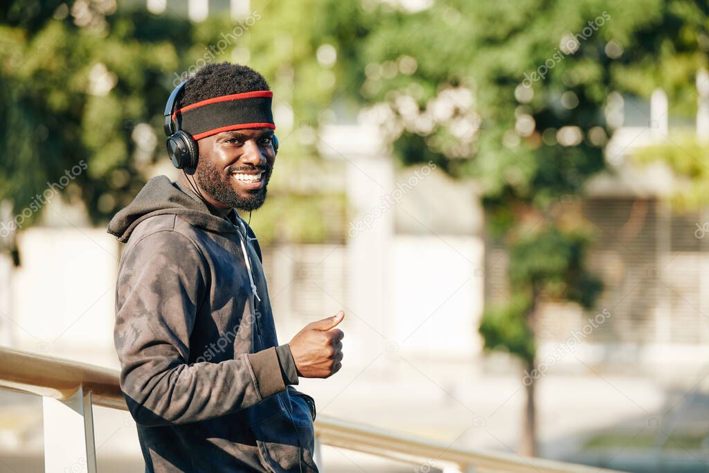 Portrait of positive young Black man listening to music in his headphones and showing thumbs-up