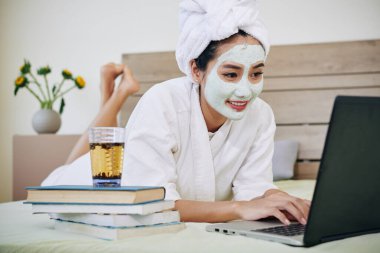 Neşeli kız öğrenci bir bardak yeşil çayla yatakta uzanıyor ve kitap yığınının üzerinde Webinar izliyor.