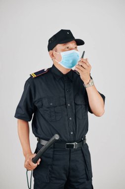 Tıbbi maskeli bir polis memuru copu tutuyor ve telsizle iş arkadaşıyla konuşuyor.