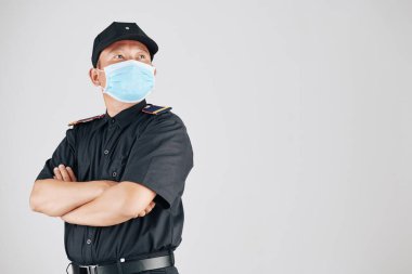 Tıbbi maskeli, kendine güvenen bir polis memuru kollarını kavuşturup, gri alana bakıyor.