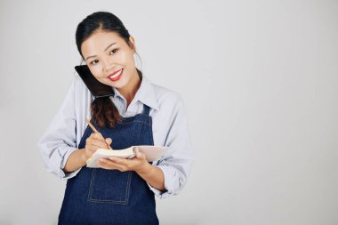 Kot önlüklü güzel Vietnamlı garson müşteriyle telefonda konuşuyor ve siparişi yazıyor.