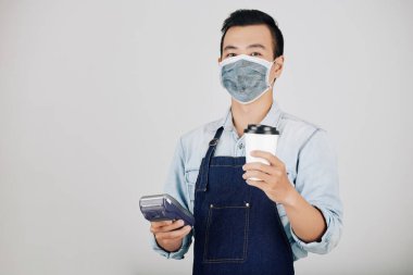 Elinde bir fincan kahve ve ödeme terminali tutan tıbbi maskeli pozitif barista.