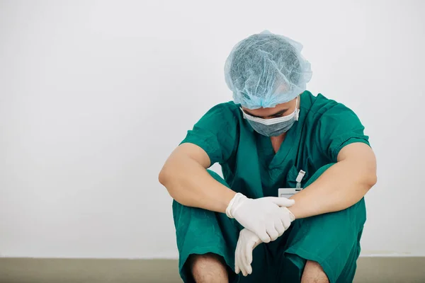 长期复杂手术后 穿着洗涤剂 橡胶手套和医用面罩的疲惫外科医生坐在地板上 — 图库照片