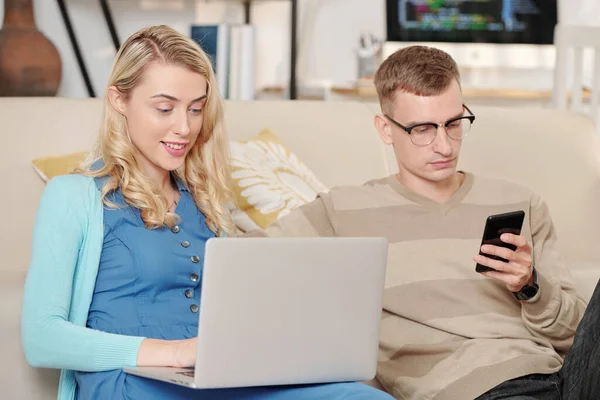 笑着坐在家里沙发上的年轻女人 在丈夫用智能手机查看社交媒体时回复同事和客户的电子邮件 — 图库照片