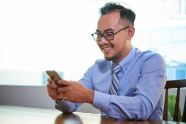 Neşeli Asyalı yönetici tek başına ofis masasında oturmuş akıllı telefonunu kullanarak mesajlar yolluyor.