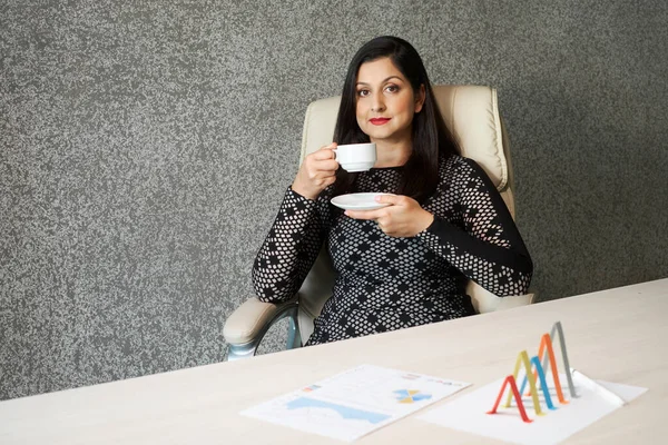 Confident mature Indian woman sitting relaxed at office desk drinking hot coffee, horizontal high angle portrait