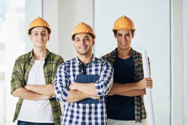 Kollarını kavuşturup kameraya gülümseyen üç profesyonel mühendisin tişört ve şapka taktığı yatay bir görüntü.