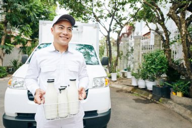 Beyaz üniformalı bir teslimatçının yatay portresi. Elinde süt şişeleri, fotokopi uzayı olan minibüsün karşısında duruyor.