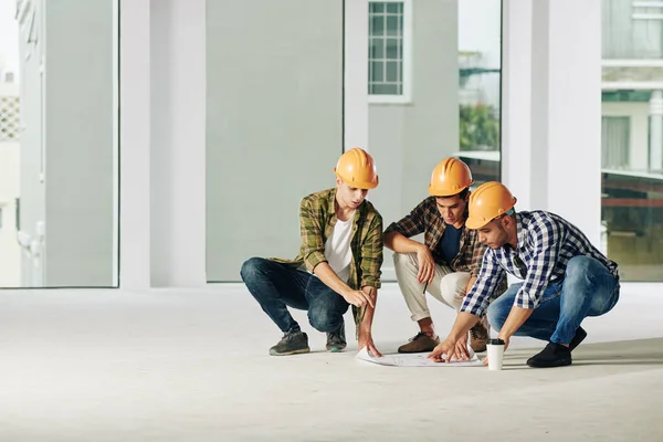 建物の計画 コピースペースを議論ハンターに座ってカジュアルな服やハード帽子を身に着けている3人の現代的な建設エンジニアのグループ — ストック写真