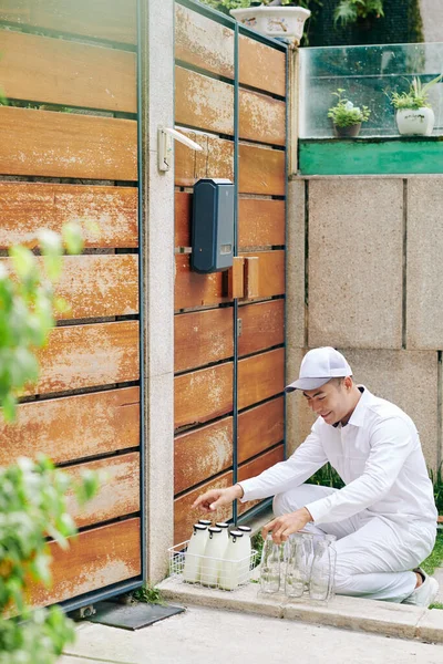 Young milkman in white uniform delivering milk and taking back empty glass bottles in morning, vertical long shot, copy space