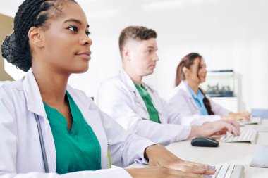 Genç siyahi pratisyen hekim hastayı ziyaret ettikten sonra bilgisayarda form dolduruyor, meslektaşları da yakınlardaki bilgisayarlarda çalışıyor.