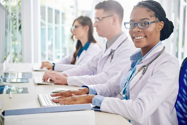穿着护目镜在电脑上工作的年轻的黑人医学研究人员笑着 她的同事在附近检查测试结果 — 图库照片