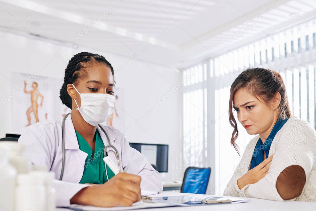 Frowning young woman suffering from pain in her chest looking at general practitioner, filling documents and writing down diagnosis and prescription