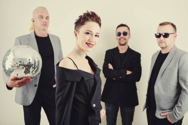 Gülümseyen çekici bayan şarkıcı kameraya bakıyor, güneş gözlüklü bando oyuncuları arka planda duruyor.