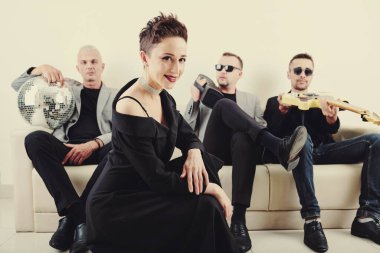 Uzun siyah elbiseli, parlak küpeli çekici bayan şarkıcının portresi, arka plandaki kanepede dinlenen müzik grubu üyeleri.