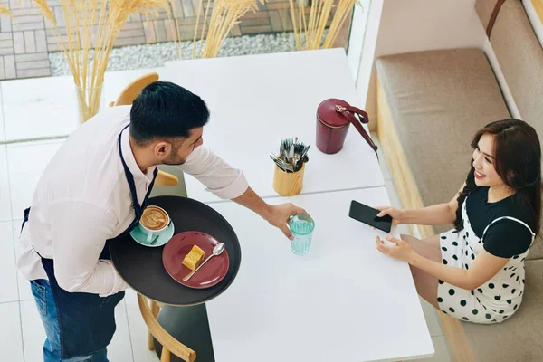 Waiter bringing glass of water, cup of cappuccino and piece of cake to pretty smiling customer, view from above