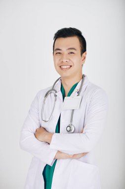 Beyaz işçi kıyafetli gülümseyen Asyalı pratisyen doktorun portresi. Kollarını kavuşturup kameraya bakıyor.