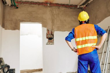 Turuncu yelek giyen ve kasketli bir adam oda duvarlarına bakıyor ve iş kapsamını, arkadan görüşü değerlendiriyor.