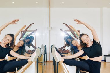 Güzel gülümseyen genç dansçılar bacak egzersizi yaparken ayaklarını bara koyuyorlar.